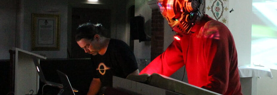 two musicians are concentrating on their laptops and controllers in what looks like a church. one of the musicians wears a sci-fi robot head.