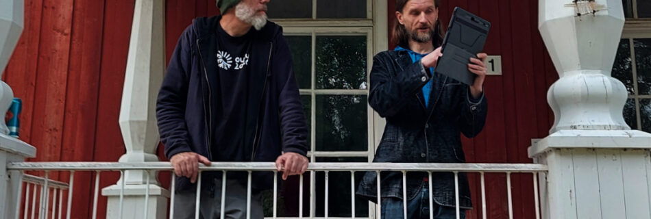 two men stand on the terrace outside an old wooden building. one is demonstrating something on an iPad.