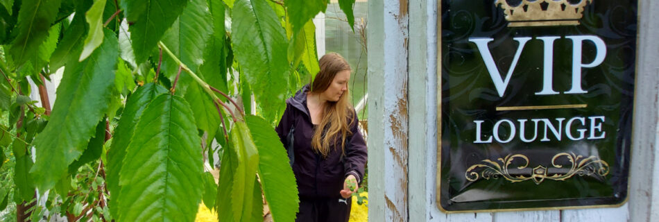 A woman in wellies stands in a greenhouse, surrounded by foliage. a sign on the wall reads: VIP LOUNGE