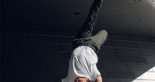 A man in casual sports clothing is doing a handstand on the concrete outside of a steel-clad building. His body is twisted into an asymmetric pose, suggesting that this is a split second of a complex breakdance choreography.