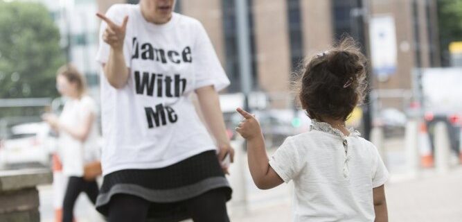 a woman and a child are dancing together on an urban street. The woman wears a t-shirt with the words DANCE WITH ME. she seems to be copying the movement of the small girl.