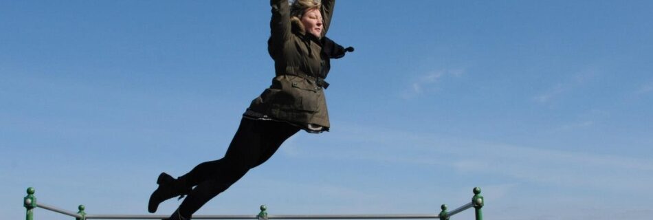 Siri is jumping, her arms outstretched above her head. Its almost a classic ballet pose, apart from the fact that she is in midair above a concrete pier overlooking the sea. There are beaches and mountains in the distance. The sky is blue, but it looks quite cold because she is wearing a coat and scarf.