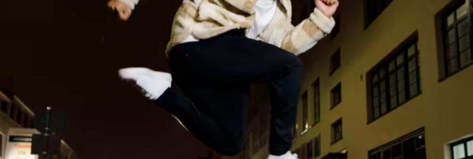 A man launches himself into the air over a night-time city street. His bright white trainers contrast with the dark background.