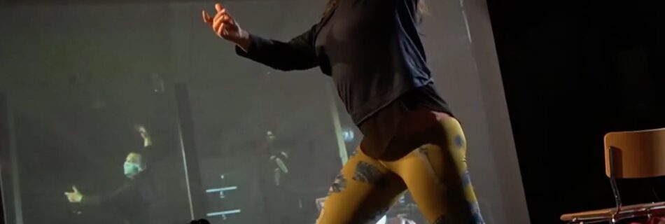 A woman is dancing with her reflection on a large screen. Also part of the image on the screen are other dancers who may or may not be in the same room.