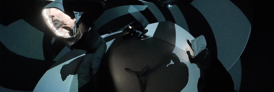 A distorted image of dancers in a dark space. There are lots of geometric blocks of light and the dancers' faces are illuminated by LED strips inside the hood of their costumes.