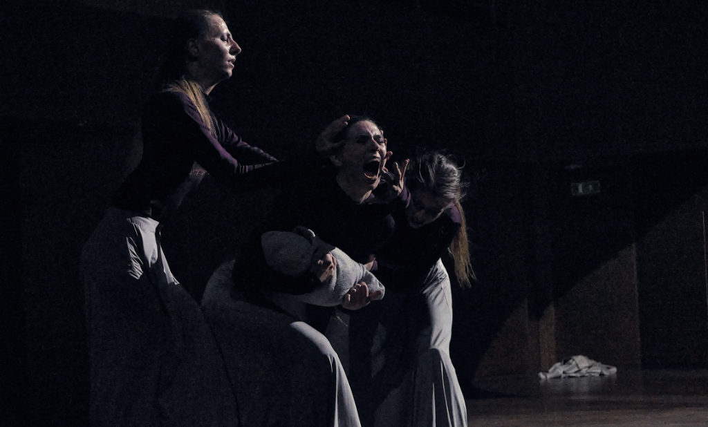 Three women are performing on stage. The scene is dark. One carries a bundle that resembles a baby. She appears to be screaming. The other two are placing their hands around her face, fingers splayed, and the effect seems to amplify her emotions.