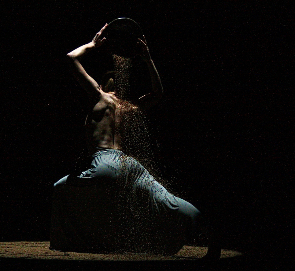 Nina is on stage, pouring some kind of grains over her back from a large bowl. The scene is quite dark, lit only by a spotlight from above. This gives it a mysterious quality and emphasises the muscular quality of her bare back and arms.