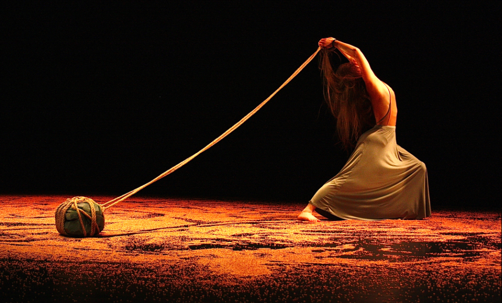 A woman in a long skirt is performing on stage. The floor is covered in some kind of grains - maybe sand or wheat. She holds a rope above her head, which trails out behind her. Tied firmly to the end of the rope is a heavy boulder - about head size - that sits on the grains strewn across the floor. The lighting is richly golden, like warm sunlight.
