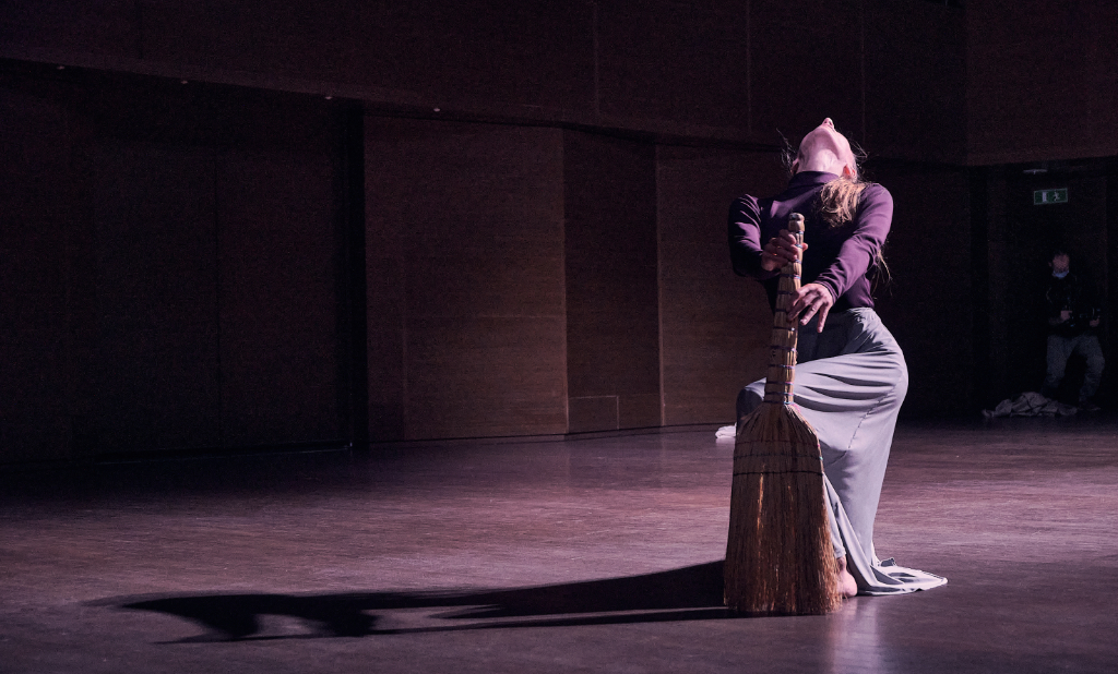 Nina dances on stage with a broom. This broom looks like an old-fashioned, traditional broom made of bound straw. She seems to be sweeping, but with exaggerated gestures, her head thrown back and the broom tentatively held in her fingertips as if it has a life of its own.